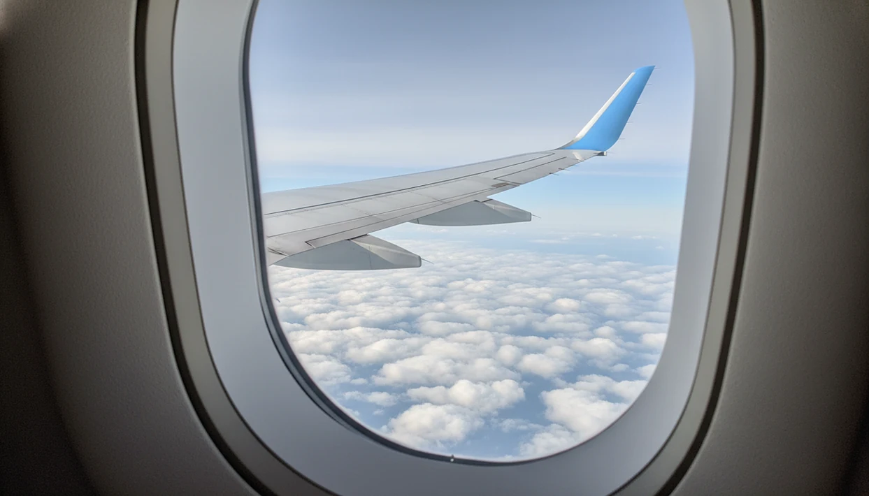 A calm passenger-window view shows an airplane wing banking gently above soft clouds in daylight.